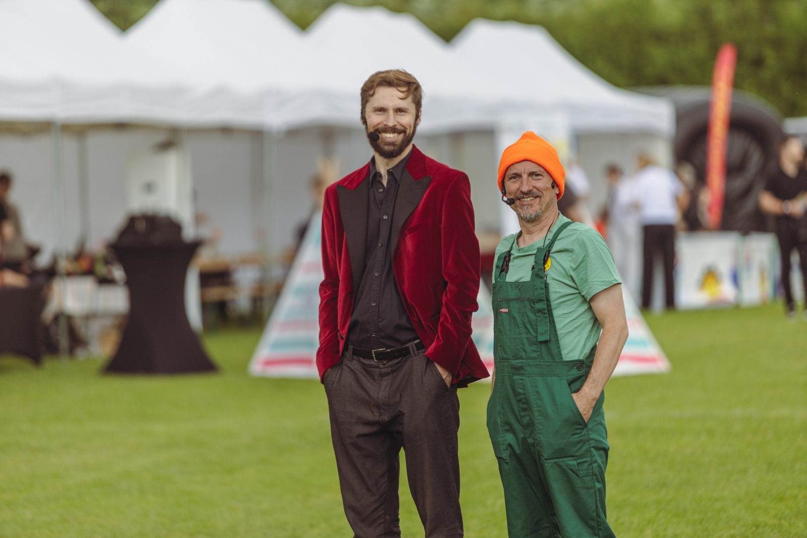 Magik w czerwonej aksamitnej marynarce i Mistrz Wpadek w pomarańczowej czapce oraz zielonych ogrodniczkach stoją na trawie przed białymi namiotami festiwalowymi.